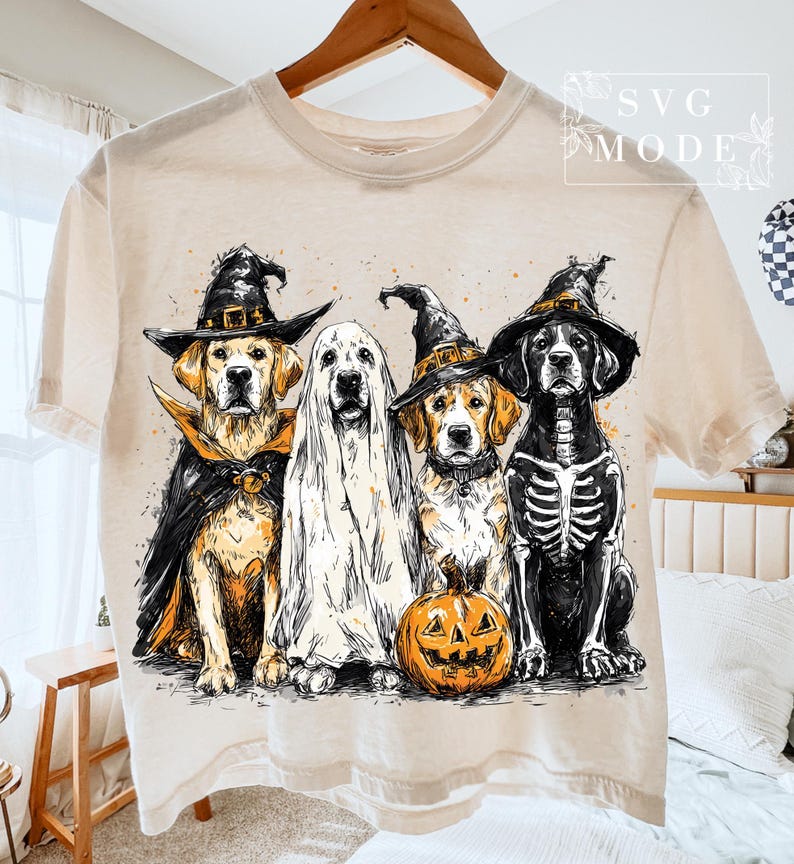 May include: Beige t-shirt featuring a Halloween-themed graphic of four dogs dressed in costumes. One dog is a vampire, another a ghost, a third a witch, and the last a skeleton. An orange pumpkin is in front of the witch dog.