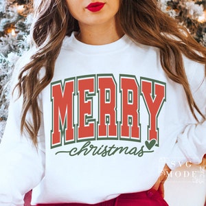 May include: White sweatshirt with the text "MERRY" in red block letters with a green outline. Below, the word "christmas" is written in a cursive font, with a green heart. The sweatshirt is a festive holiday design.