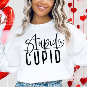 May include: White sweatshirt with the words "Stupid Cupid" in black script and block letters. The word "Cupid" is underlined with a heart symbol. The sweatshirt is a Valentine's Day theme.