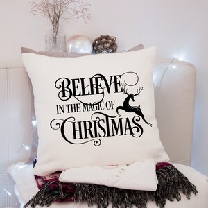 Puede incluir: Coj&iacute;n cuadrado color crema con las palabras "BELIEVE IN THE MAGIC OF CHRISTMAS" en escritura negra y una silueta de reno negro. El coj&iacute;n est&aacute; sobre una superficie blanca con una camisa blanca y una bufanda con flecos.