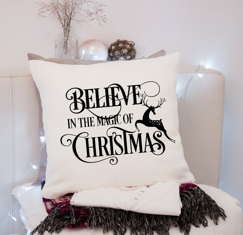 Puede incluir: Coj&iacute;n cuadrado color crema con las palabras "BELIEVE IN THE MAGIC OF CHRISTMAS" en escritura negra y una silueta de reno negro. El coj&iacute;n est&aacute; sobre una superficie blanca con una camisa blanca y una bufanda con flecos.
