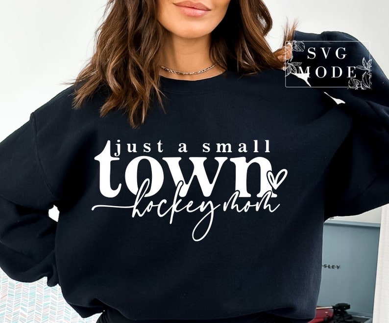 Op de afbeelding: Zwart sweatshirt met witte tekst die "Just a small town hockey mom" zegt met een klein hartje.
