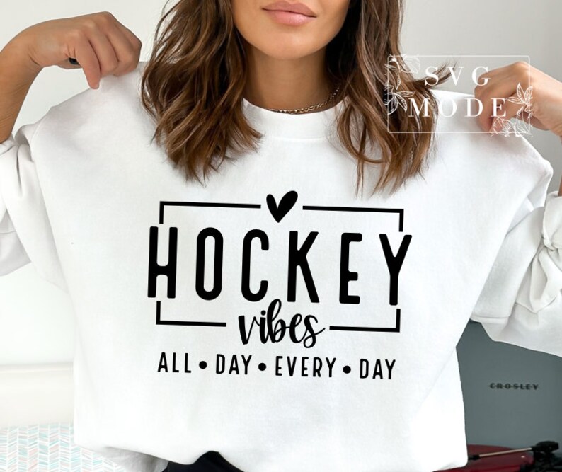 Puede incluir: Sudadera blanca con texto negro que dice "Hockey vibes all day every day" con un coraz&oacute;n sobre la palabra "Hockey".