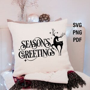 May include: White pillow with black text that reads "Season's Greetings" and a black silhouette of a deer with antlers. The pillow is on a white surface with a string of lights.
