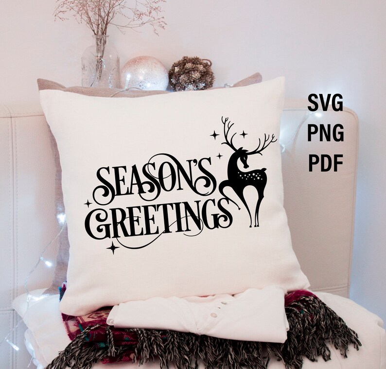 May include: White pillow with black text that reads "Season's Greetings" and a black silhouette of a deer with antlers. The pillow is on a white surface with a string of lights.