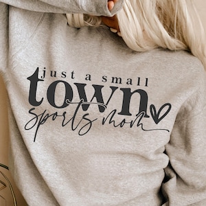 Pode incluir: Um moletom cinza claro com o texto "just a small town sports mom" em letras pretas e em bloco. O moletom tem gola redonda e mangas compridas.