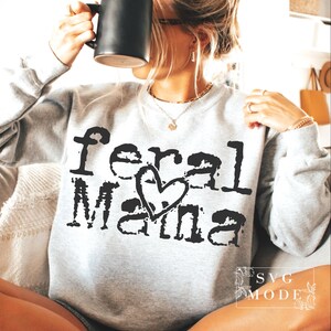 Könnte beinhalten: Hellgraues Sweatshirt mit dem Text "Feral Mama" in verblichener schwarzer Schrift und einem Herz-Grafik. Das Sweatshirt hat einen Rundhalsausschnitt. Das Bild zeigt auch eine schwarze Tasse.