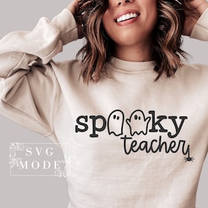 May include: A beige sweatshirt with the text "spooky teacher" in black lettering. Two ghost silhouettes are included in the text. A small black spider is in the bottom right corner.