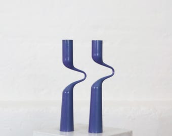 May include: Two matching purple candle holders featuring a smooth, solid finish. Each piece has a cylindrical base that transitions into a distinct, curved, S-shaped middle section, leading up to a straight, vertical top opening for a candle.