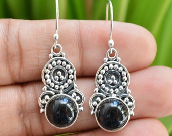 May include: A pair of silver-toned dangle earrings. Each earring features a dark, round gemstone set within a decorative, circular frame with small beaded accents. The earrings are suspended from a simple hook.