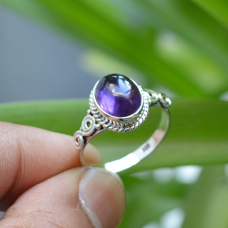 May include: A silver ring with an oval amethyst gemstone. The ring has a delicate, ornate design with swirls and a beaded edge.