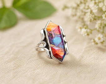 Puede incluir: Un anillo de plata con una piedra larga de corte hexagonal que presenta un patrón multicolor vibrante en tonos naranja, morado y azul brillante. El engaste de metal pulido tiene aberturas decorativas curvas a los lados del aro.
