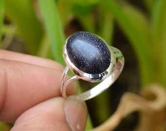 Puede incluir: Un anillo de plata con una gran piedra preciosa ovalada de arenisca azul con inclusiones brillantes. El anillo tiene un diseño sencillo con una caña dividida. La piedra preciosa está engastada en un bisel pulido, mostrando el color y la textura de la piedra.