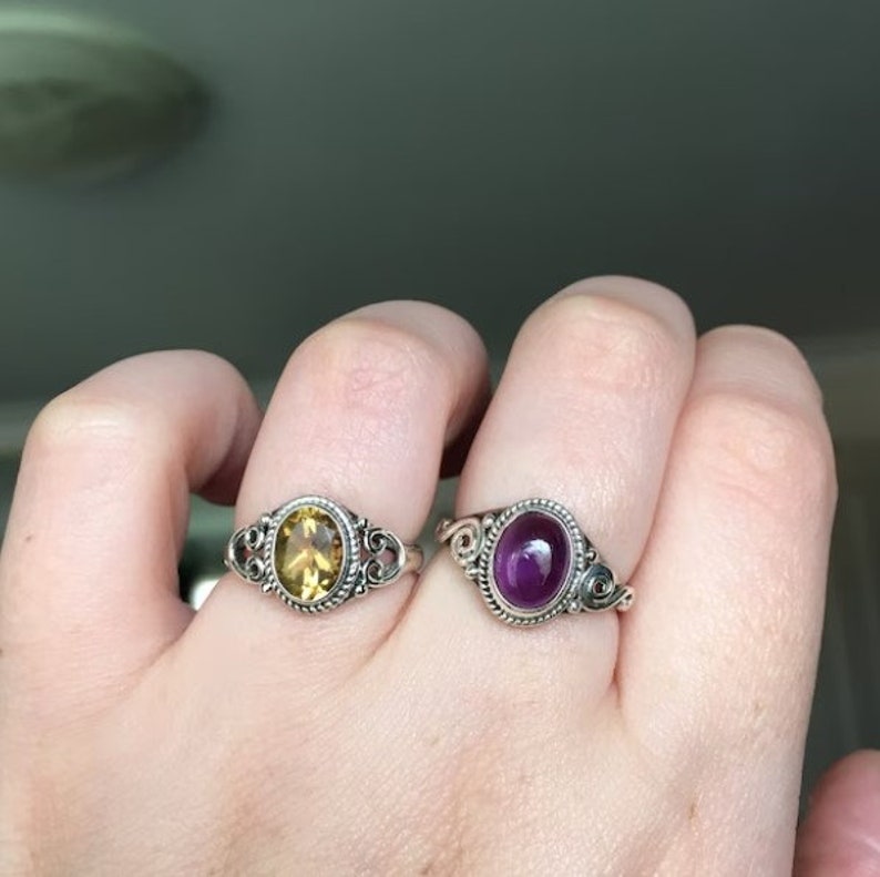May include: Two silver rings with oval-shaped gemstones. The ring on the left has a yellow gemstone, and the ring on the right has a purple gemstone.