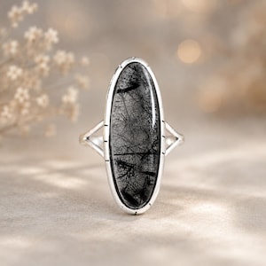 Puede incluir: Un anillo de plata con una gran piedra preciosa de cuarzo rutilado negro de forma ovalada y alargada. La piedra está engastada en un bisel de plata liso con pequeñas muescas decorativas a lo largo del borde. El aro se divide en dos hilos finos.