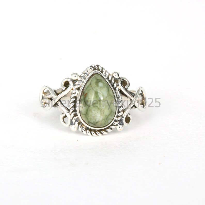 May include: A silver ring with a green gemstone set in a teardrop shape. The ring has a detailed design with intricate swirls and a rope-like border around the stone.