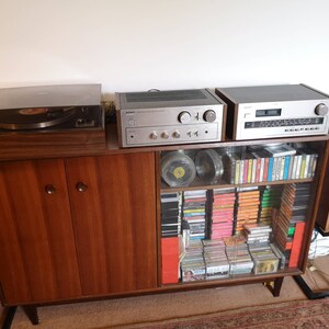 Puede incluir: Un sistema de sonido est&eacute;reo vintage con un tocadiscos, un amplificador y altavoces. El sistema est&aacute; instalado en un gabinete de madera con estantes de vidrio llenos de cintas de cassette y CD. Los altavoces est&aacute;n en soportes negros.