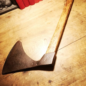 Puede incluir: Un hacha antigua con un mango de madera desgastada y una hoja de metal oscura y envejecida. El hacha está colocada en diagonal sobre una superficie de madera, sugiriendo un tema rústico o histórico. Libros rojos son visibles en el fondo.