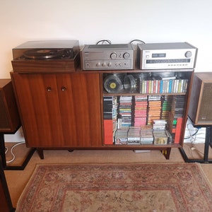 Puede incluir: Un sistema de sonido est&eacute;reo vintage con un tocadiscos, un amplificador y altavoces. El sistema est&aacute; instalado en un gabinete de madera con estantes de vidrio llenos de cintas de cassette y CD.