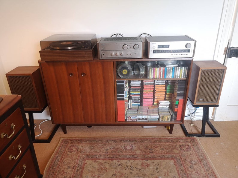 Puede incluir: Un sistema de sonido est&eacute;reo vintage con un tocadiscos, un amplificador y altavoces. El sistema est&aacute; instalado en un gabinete de madera con estantes de vidrio llenos de cintas de cassette y CD.