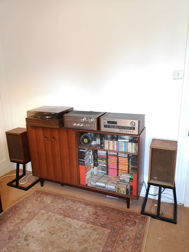 Puede incluir: Un sistema de sonido est&eacute;reo vintage con un tocadiscos, un amplificador y altavoces. El sistema est&aacute; instalado en un gabinete de madera con un estante de vidrio lleno de cintas de cassette y CD.