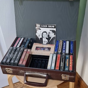 Può includere: Una valigetta di trasporto in pelle marrone con maniglia e chiusura. La valigetta è aperta e piena di cassette audio. Le cassette sono etichettate con vari titoli, tra cui "The Goon Show", "Earth Wind & Fire", "Time For Old Time", "Right Said Fred Up" e "Star Power".