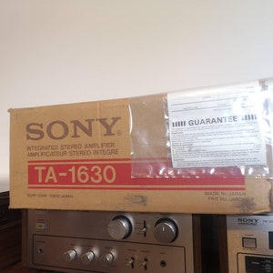 Puede incluir: Una caja de cart&oacute;n marr&oacute;n con las palabras "SONY" y "TA-1630" impresas en ella. La caja es para un amplificador est&eacute;reo integrado. La caja est&aacute; encima de un receptor est&eacute;reo plateado con una placa frontal negra y un sintonizador plateado.