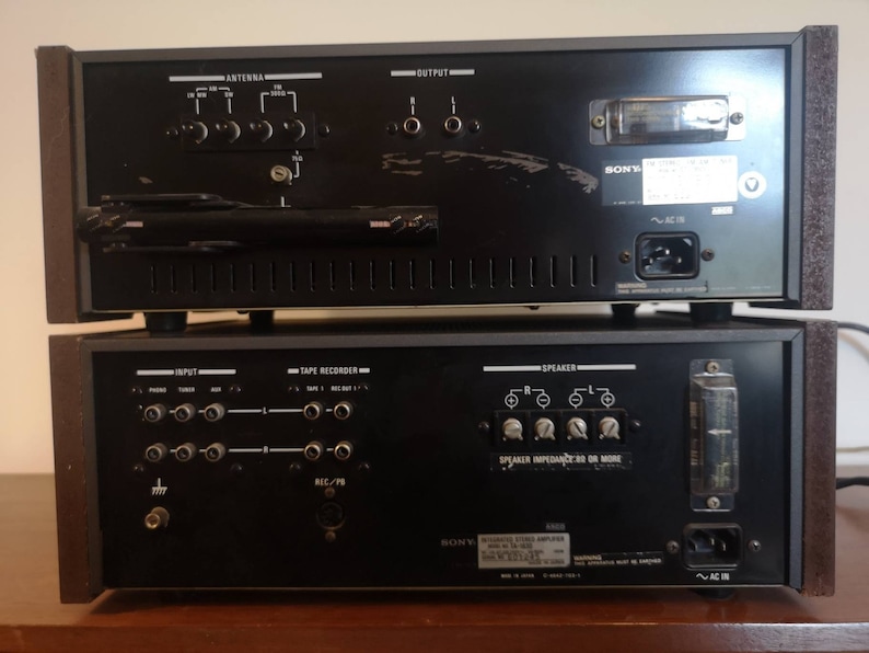 Puede incluir: Vista trasera de un receptor est&eacute;reo y un amplificador Sony vintage. El receptor tiene una cara de metal negro con una perilla plateada y una antena de pl&aacute;stico negro. El amplificador tiene una cara de metal negro con perillas plateadas y un cable de alimentaci&oacute;n de pl&aacute;stico negro.
