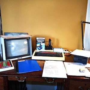 May include: A vintage computer setup with a black and white monitor, a keyboard, a joystick, and a floppy disk drive. The computer is on a wooden desk with a blue folder, papers, and a lamp.