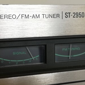 Puede incluir: Primer plano de un sintonizador est&eacute;reo plateado con una placa frontal negra. La placa frontal tiene dos medidores anal&oacute;gicos, uno etiquetado como "SIGNAL" y el otro como "FM TUNING". El sintonizador est&aacute; etiquetado como "M STEREO/FM-AM TUNER | ST-2950F".