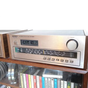 Puede incluir: Un sintonizador FM est&eacute;reo/AM Sony ST-295F plateado con un panel lateral de madera. El sintonizador tiene un dial plateado con n&uacute;meros negros y una perilla de sintonizaci&oacute;n plateada.