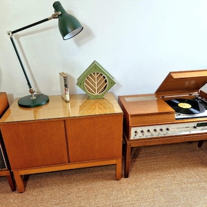 May include: A vintage entertainment unit featuring a record player, speakers, and a cabinet. A green desk lamp and a decorative green and wood accent piece are on the cabinet. The record player has a black vinyl record on the turntable.