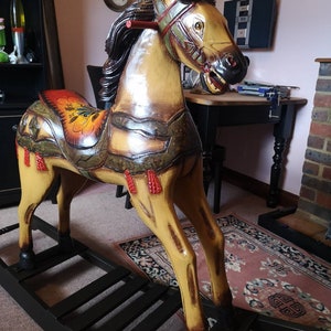 May include: A wooden rocking horse with a yellow, brown and green painted finish. The horse has a saddle with a red, yellow and orange floral design. The horse is on a black wooden rocking base.