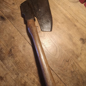 Puede incluir: Un hacha antigua con una hoja de acero oscura y desgastada y un mango de madera. La cabeza del hacha es rectangular con un filo curvo. El mango es de color marrón claro y parece desgastado. El hacha está sobre una superficie de madera.