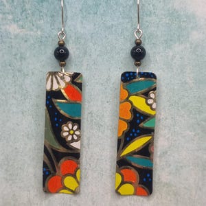 Pode incluir: Um par de brincos retangulares com fundo preto e um padrão floral vibrante em laranja, amarelo, turquesa e branco. Cada brinco pende de um gancho prateado e inclui uma conta preta acima da peça retangular.