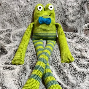 May include: A green crocheted frog with a blue bow tie sitting on a gray fuzzy blanket. The frog has long green arms and legs with gray and green stripes.