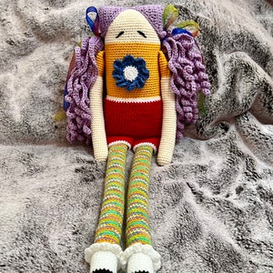 Puede incluir: Una muñeca de crochet con pelo de lana morado, un vestido amarillo y rojo y una flor azul en el pecho. La muñeca está sentada sobre una superficie gris y peluda.