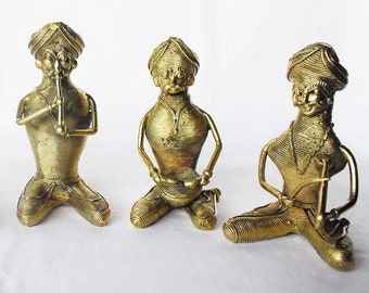 May include: Three gold wire sculptures of musicians playing instruments. The musicians are wearing traditional Indian clothing and turbans. The sculptures are sitting on a white surface.