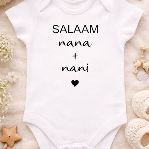 May include: A white baby bodysuit featuring the text "SALAAM nana + nani" in black, with a small black heart. The garment is displayed on a soft, white surface with decorative items.