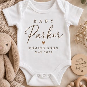 May include: A white short-sleeved baby bodysuit featuring brown text that reads BABY Parker, a small heart icon, and the text COMING SOON MAY 2027. The garment has a round neckline and three metal snaps at the bottom for closure.