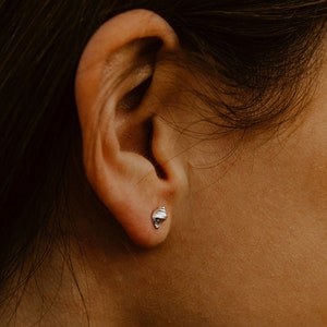 May include: A silver stud earring is visible on a person's ear. The earring features a small, detailed design. The person's dark brown hair and blurred background create a natural, earthy aesthetic.