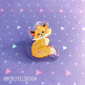 May include: A clear acrylic charm featuring a cartoon fox with orange fur, a white chest, and a purple crescent moon and stars on its tail. The fox is smiling and has closed eyes.
