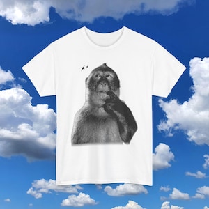 May include: White t-shirt featuring a black and white illustration of a monkey with its finger to its mouth, as if deep in thought. Two small black birds are above the monkey's head. The shirt is set against a blue sky with white clouds.