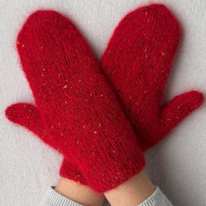 Puede incluir: Un par de manoplas de punto de color rojo brillante con una superficie texturizada y difusa, con pequeñas motas multicolores tejidas en el hilo. Las manoplas tienen una forma simple y redondeada y se muestran juntas sobre un fondo neutro.