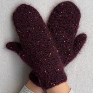 May include: A pair of knitted burgundy mittens with flecks of yellow and orange. The mittens are made of a soft, textured yarn and are shown against a light gray background. The mittens are designed to keep hands warm.