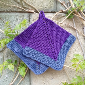 Könnte beinhalten: Zwei quadratische, gestrickte Topflappen. Die Mitte ist tiefviolett, mit einem hellblauen Rand. Eine Schlaufe zum Aufhängen ist angebracht. Die Topflappen werden vor einer strukturierten Steinmauer mit grünen Weinreben gezeigt.