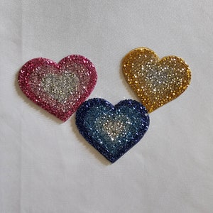 May include: Three heart-shaped patches with a sparkly, iridescent finish. The hearts are pink, blue, and gold.