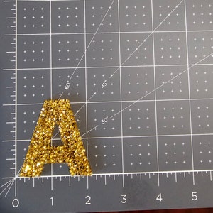 May include: A gold glitter letter A on a gray cutting mat with white markings.