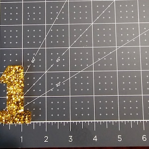 May include: A gold glitter number one, approximately 2 inches tall, on a gray cutting mat with white grid lines.