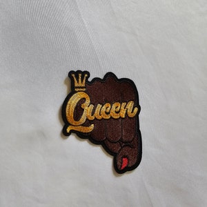 May include: A brown fist with a gold crown and the word "Queen" embroidered in gold. The fist has red nail polish.